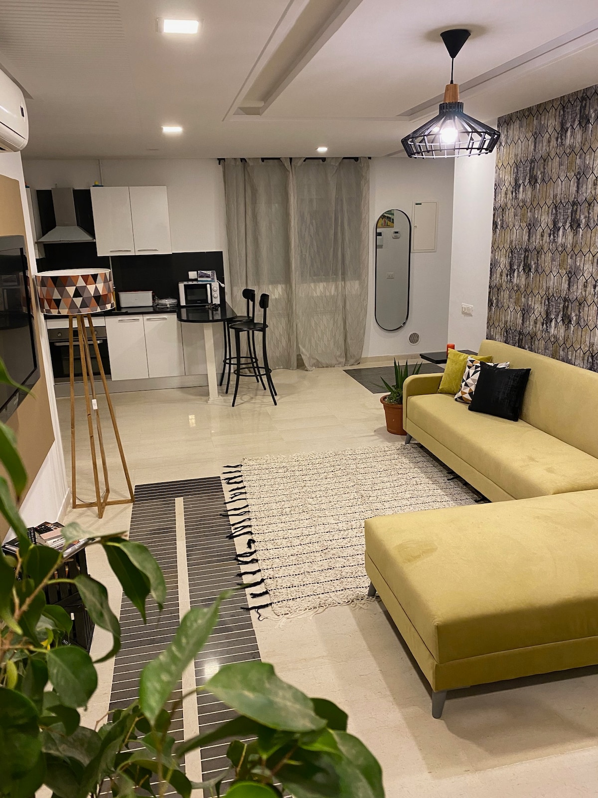 An open living area includes a modern yellow couch and a small dining table with high stools. A kitchenette is visible in the background, equipped with essential appliances. A potted plant adds a touch of greenery, while curtains frame a large window allowing natural light.
