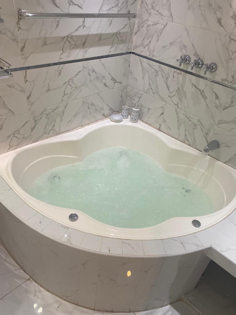 A heart-shaped jacuzzi is featured, set in a serene marble-tiled bathroom. Soft bubbles rise from the water, and decorative soap dispensers are placed nearby. The surrounding walls are lined with elegant marble patterns, contributing to an atmosphere of relaxation.