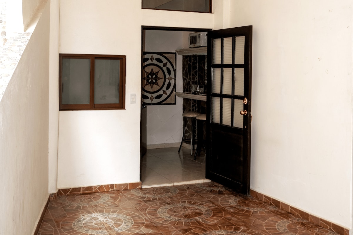 The entrance features a tiled floor with intricate patterns and a large wooden door that opens into the living space. A small window alongside the door provides natural light, and a glimpse of the interior shows a decorative wall and dining area.