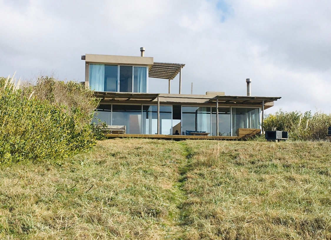 The house features a modern design with large glass windows, providing panoramic views of the surroundings. A wooden deck extends out from the building, enhancing the connection with the outdoors. The structure is set against a backdrop of lush greenery and a clear sky.