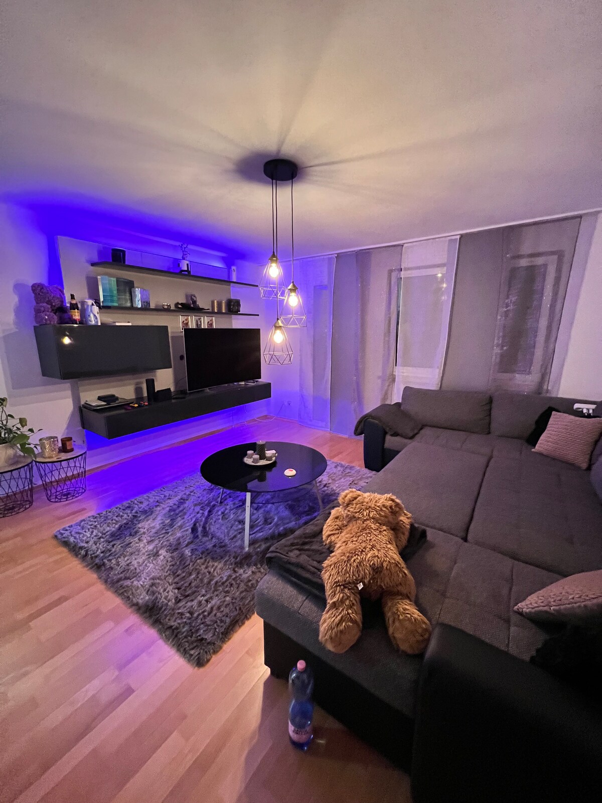 A spacious living room features a large sectional couch with a decorative throw, facing a low coffee table. Soft lighting from pendant lamps and LED accents creates a calming ambiance. A plush rug sits underfoot, while a television is mounted on the wall with shelving above.