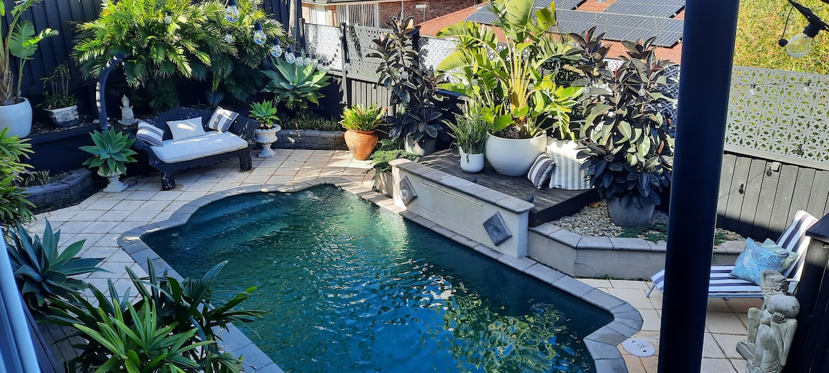 An inviting outdoor pool area is surrounded by lush tropical plants and decorative features. A comfortable lounging area is positioned nearby, with a sectional sofa and a sunbed. The pool's clear water glistens under natural light, offering a serene backdrop for relaxation.