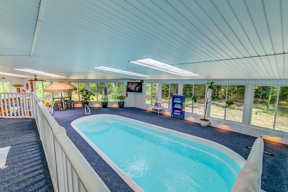 An indoor pool is surrounded by large windows, allowing natural light to fill the space. The pool area features a soft blue water, comfortable seating, and plants adding accents of greenery. A television and tropical-themed decor enhance the relaxing environment.
