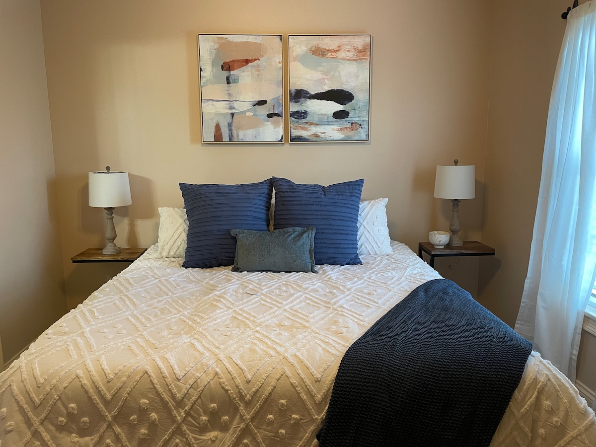 A spacious bed is centered in the room, adorned with a textured white coverlet and decorative blue pillows. Two bedside lamps with soft, warm light stand on wooden nightstands. Abstract artwork in muted tones is displayed above the bed, complementing the calming color palette.