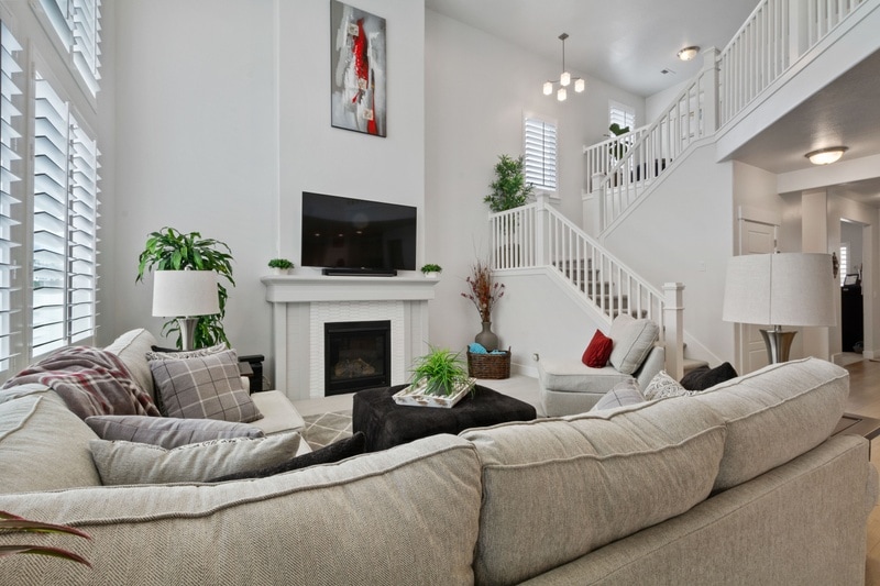 A spacious living room is defined by neutral tones and natural light, featuring a large sectional sofa adorned with various cushions. A fireplace is centered beneath a mounted TV, while modern light fixtures hang above. Elegant plants add a touch of greenery to the inviting space.