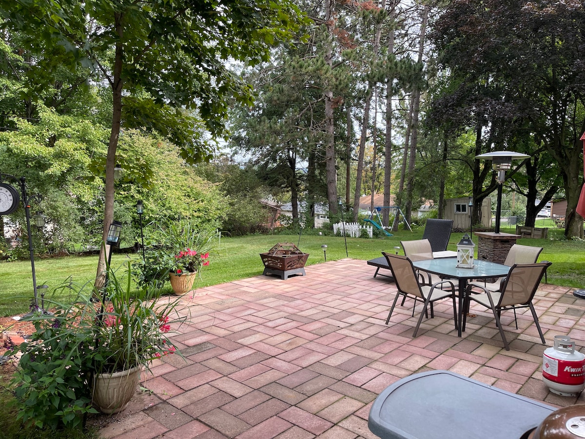 A spacious outdoor patio is surrounded by greenery, featuring a fire pit and several planters with blooming flowers. Comfortable seating is arranged around a dining table, and a heater stands in one corner, enhancing outdoor usability. The lawn stretches beyond the patio, framed by trees.