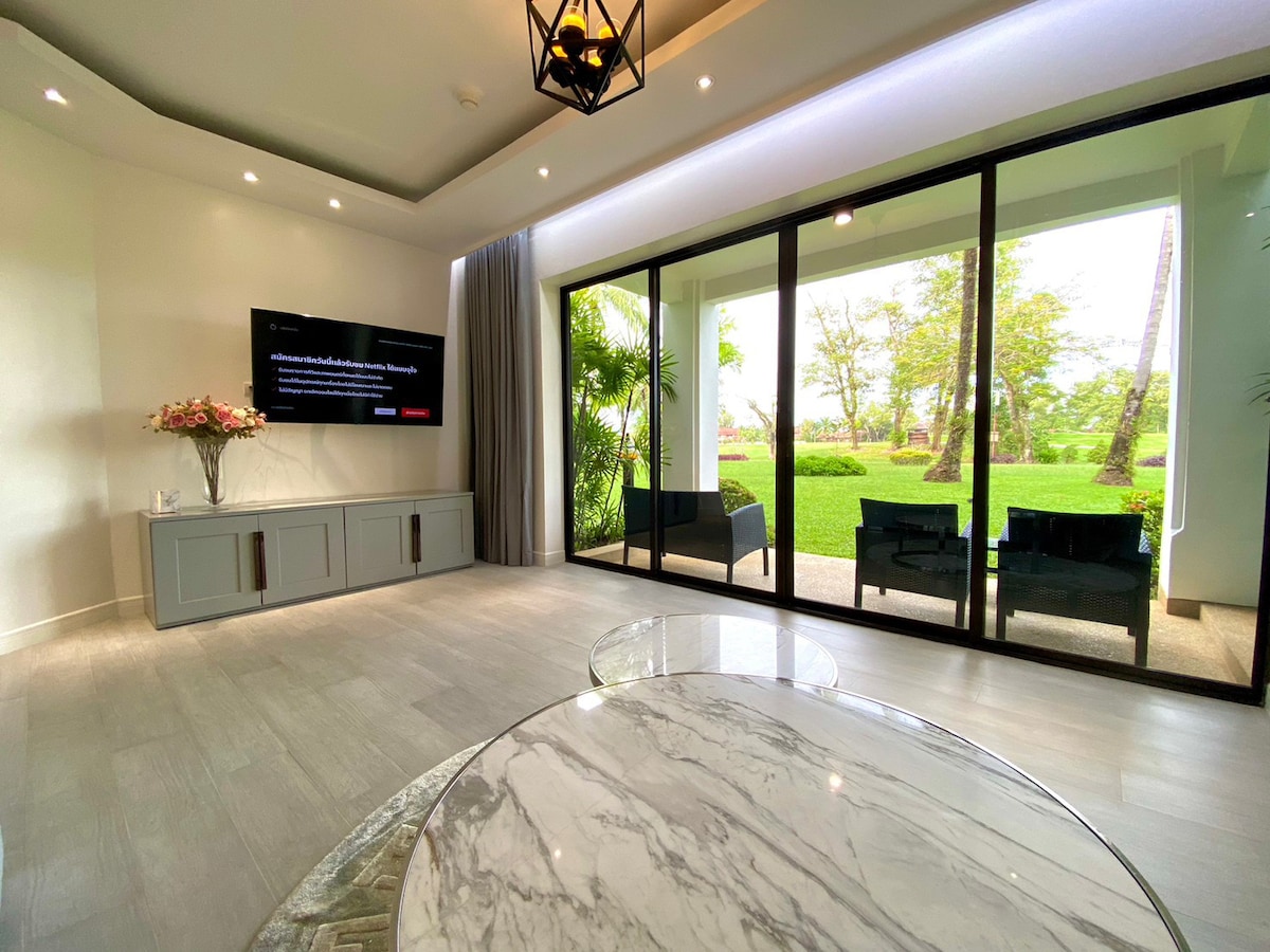 A spacious living area features a large smart TV mounted on a neutral-toned wall, complemented by a stylish marble coffee table. Floor-to-ceiling glass doors provide views of the lush garden, allowing natural light to fill the space while seamlessly connecting indoor and outdoor areas.