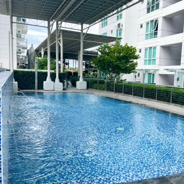 A swimming pool is set within a landscaped area, featuring clear blue water and mosaic tiles along the edge. The surrounding greenery and white structures create an inviting atmosphere. A shaded seating area is available beside the pool, offering a place to relax.