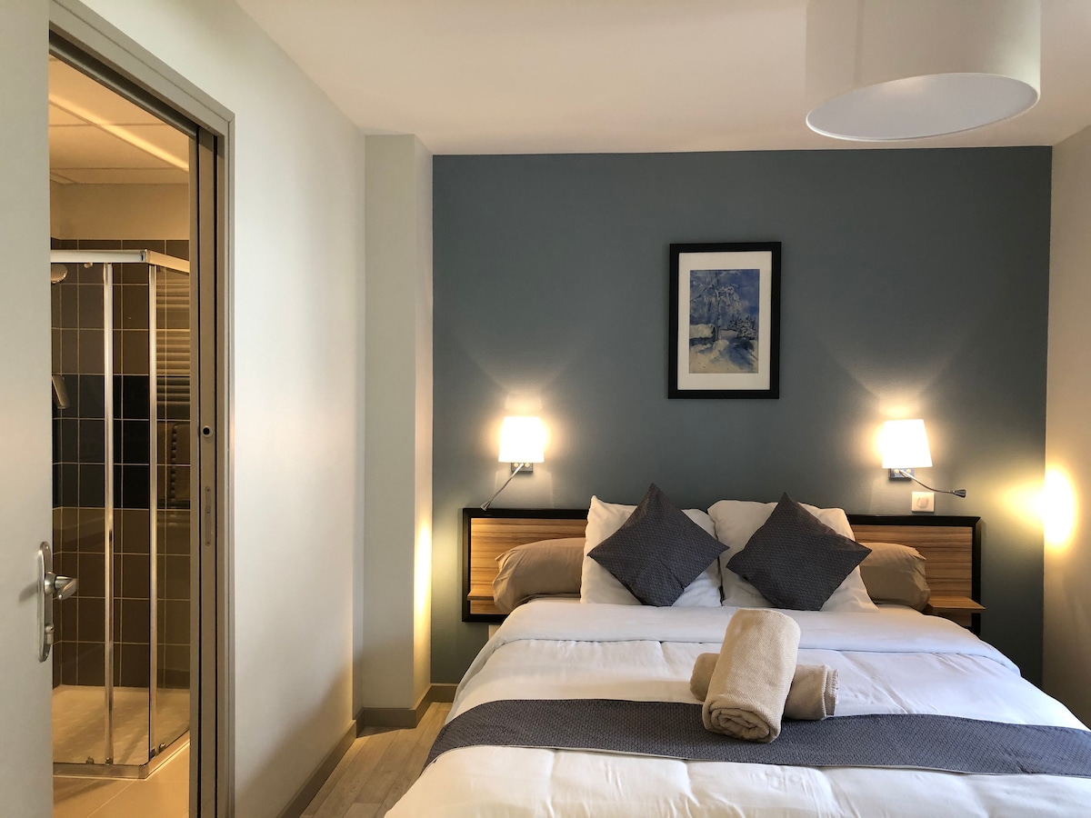 A cozy bedroom features a spacious bed with two decorative pillows, flanked by warm bedside lamps. A light blanket is neatly placed at the foot of the bed. A framed artwork adorns the wall, and an en-suite bathroom can be seen through an open door.