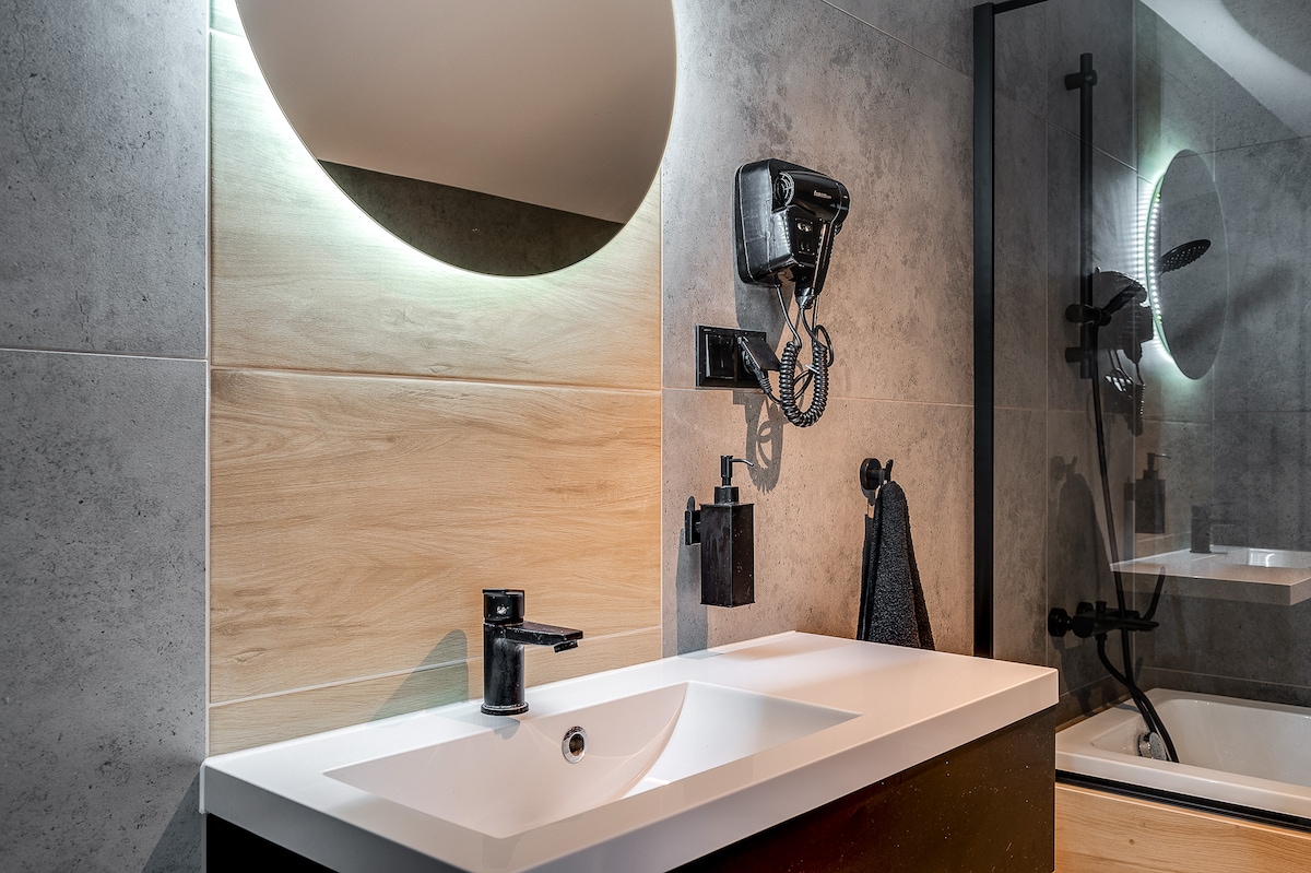 A modern bathroom is featured, showcasing a sleek countertop with a rectangular sink and a stylish faucet. A large round mirror above the sink is illuminated by soft lighting. A hairdryer is mounted on the wall, alongside a dark towel hanging nearby. A glass-enclosed shower is visible in the background.