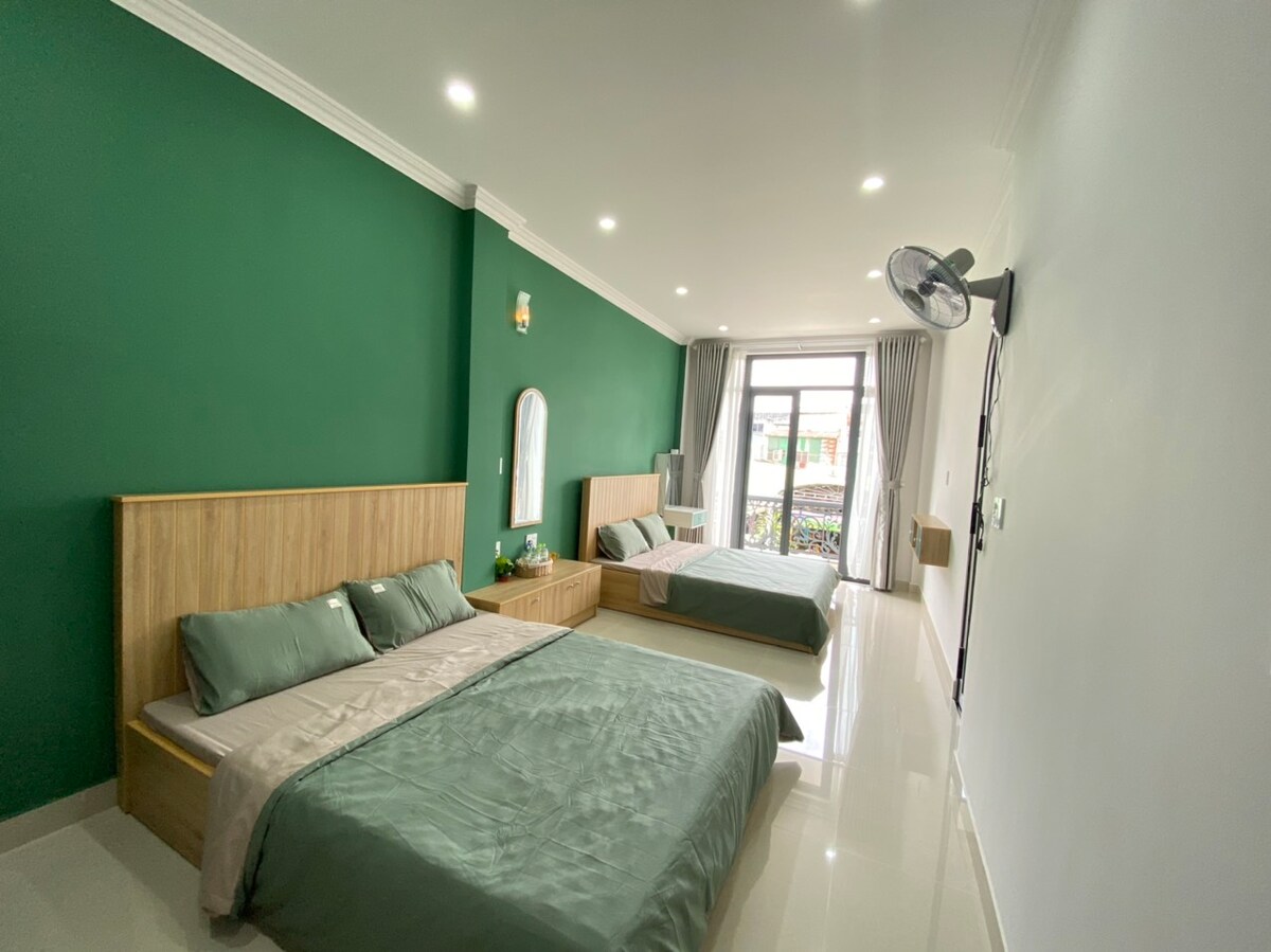 Two spacious beds are positioned in a bright room featuring green accent walls. Natural light is filtered through a large window with sheer curtains, illuminating the minimalist decor. A mirror and bedside tables are present, enhancing the room's functionality.