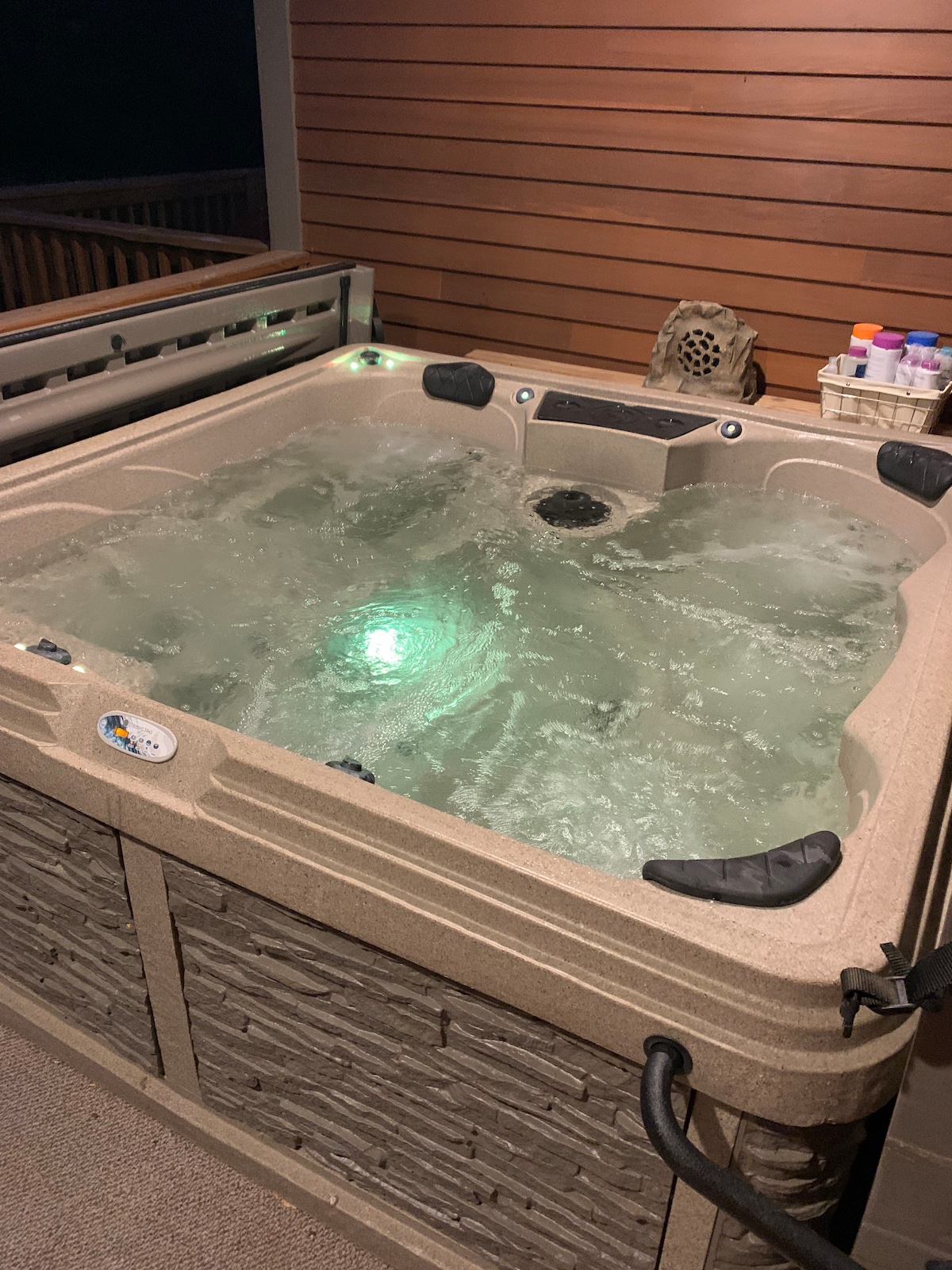 A spacious hot tub is situated on a covered deck, illuminated by soft green lighting. The tub features built-in seating and jets, surrounded by a textured exterior. Essentials are neatly arranged nearby, enhancing the overall relaxation space.