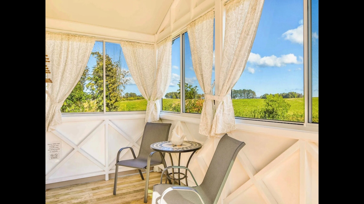 A sunlit sitting area features large windows draped with sheer curtains, offering panoramic views of the rural landscape. A small table is complemented by two comfortable chairs, creating a serene spot for relaxation. Natural light floods the space, enhancing the peaceful atmosphere.
