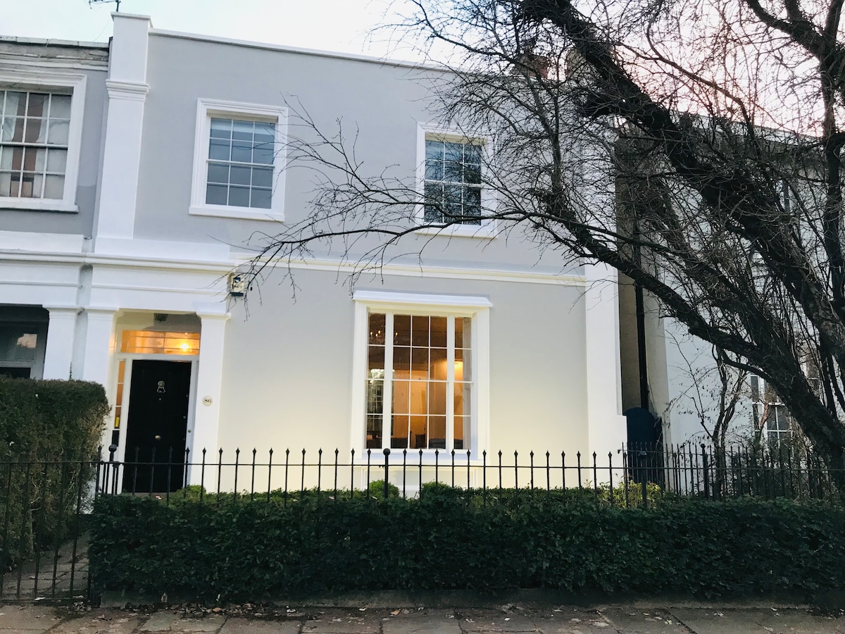 The exterior of a Georgian-style home is shown, with a symmetrical façade featuring large sash windows. A neatly manicured hedge surrounds the front garden, while a black wrought-iron fence defines the property boundaries. The structure is painted in a light grey, providing a modern touch.