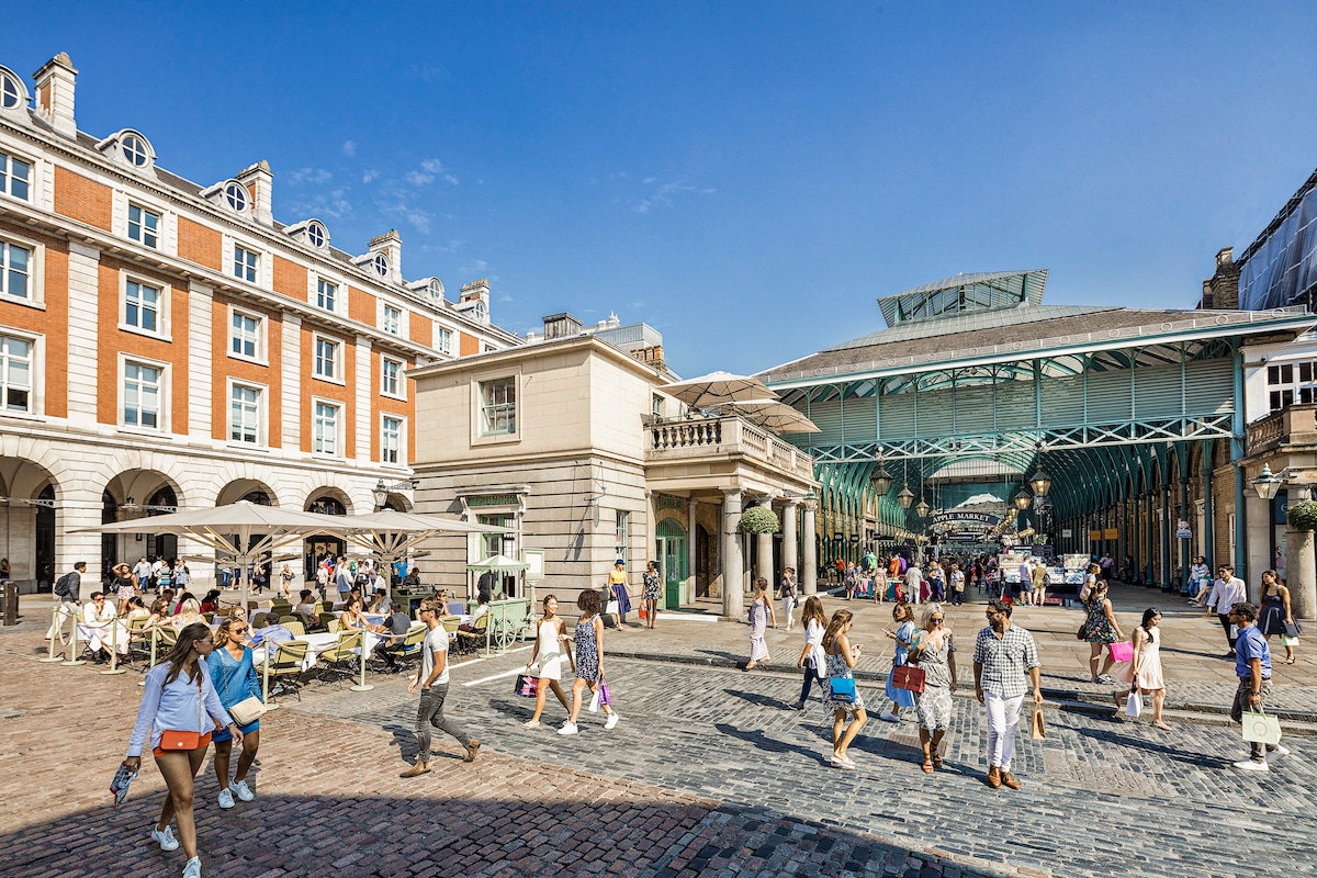 A bustling Covent Garden scene is captured, showcasing a vibrant plaza lined with shops and outdoor seating areas. Visitors stroll along cobblestone pathways, with historic buildings and a modern glass structure in the background under a clear blue sky.