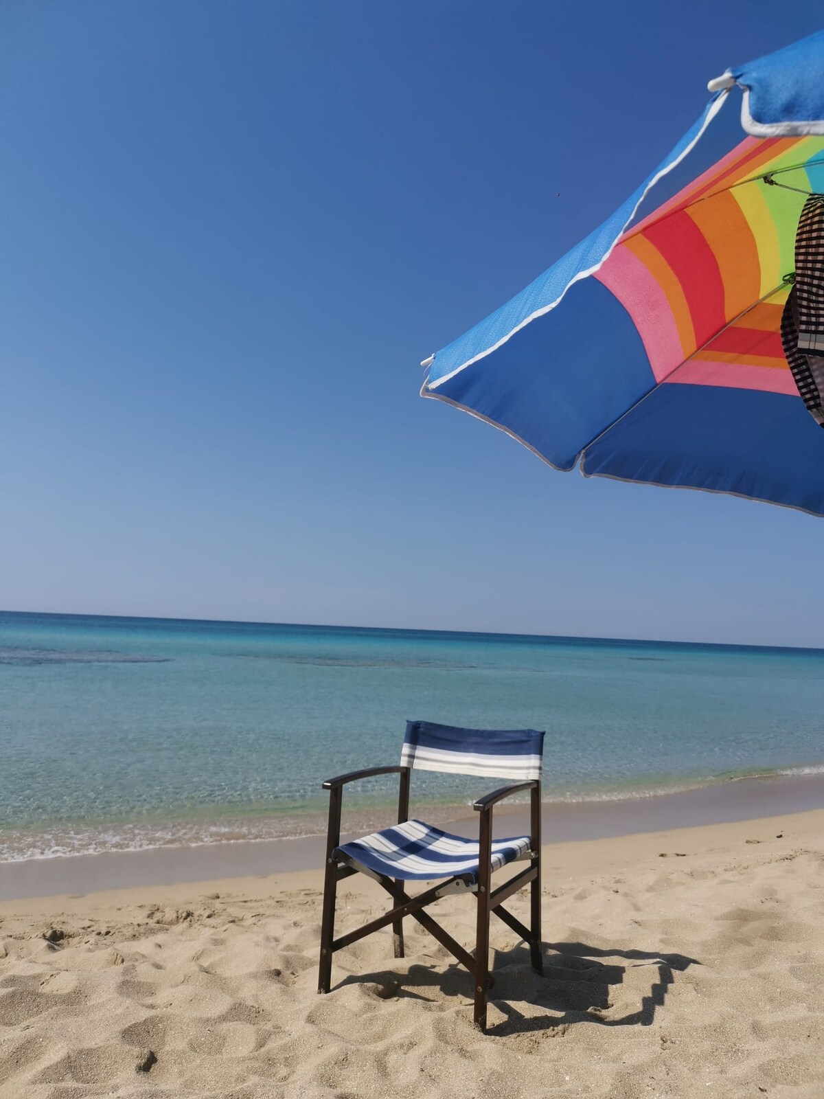 A beach scene includes a striped beach umbrella and a folding chair positioned on the sandy shore. The clear water reflects shades of blue and green, with gentle waves lapping at the sand. The sky is bright and cloudless, suggesting a calm day.