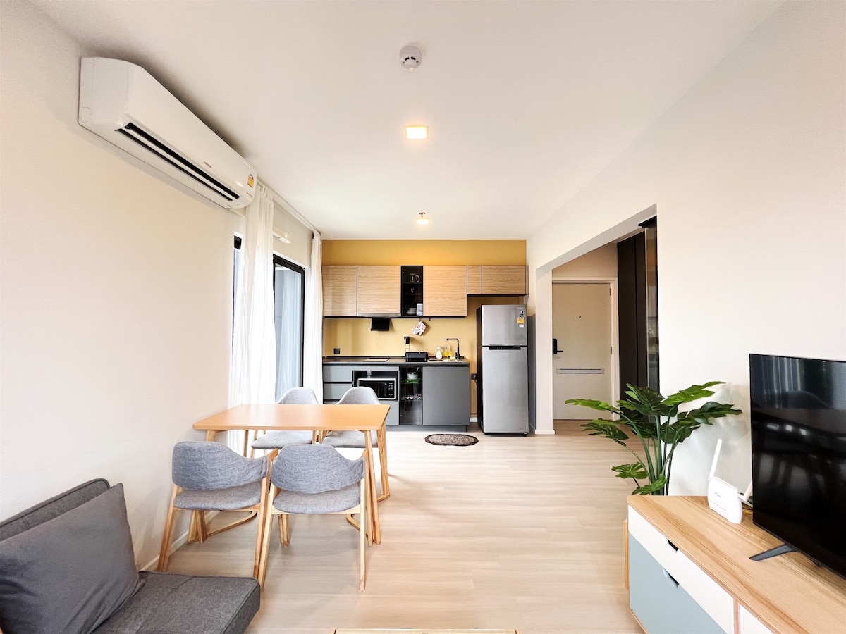 A modern living area with a comfortable sofa is accompanied by a dining table and chairs. The kitchen area features sleek cabinetry and appliances, including a refrigerator. Natural light is allowed in through large windows, while a decorative plant adds a touch of greenery.