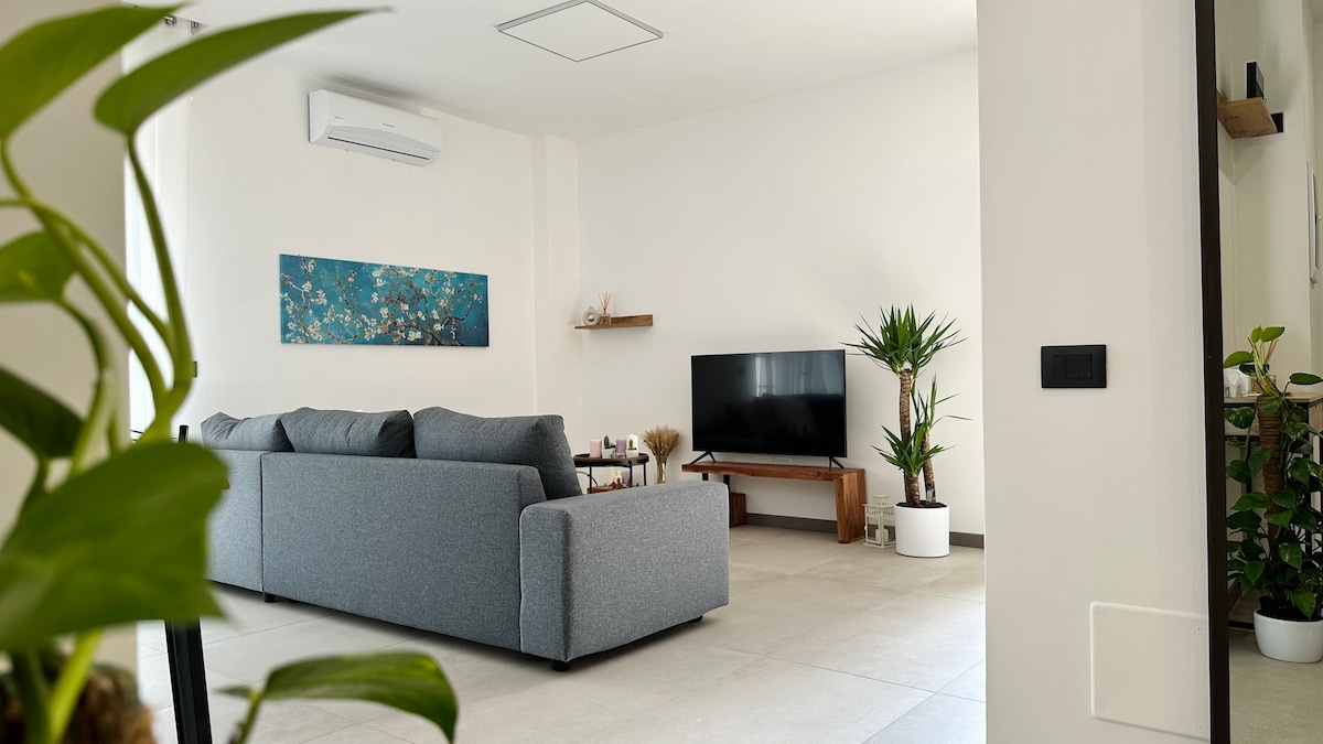 A modern living space features a gray sectional sofa positioned across from a flat-screen television. A decorative blue artwork hangs on the wall, while indoor plants add a touch of greenery. Light-colored tiles enhance the bright and airy atmosphere.
