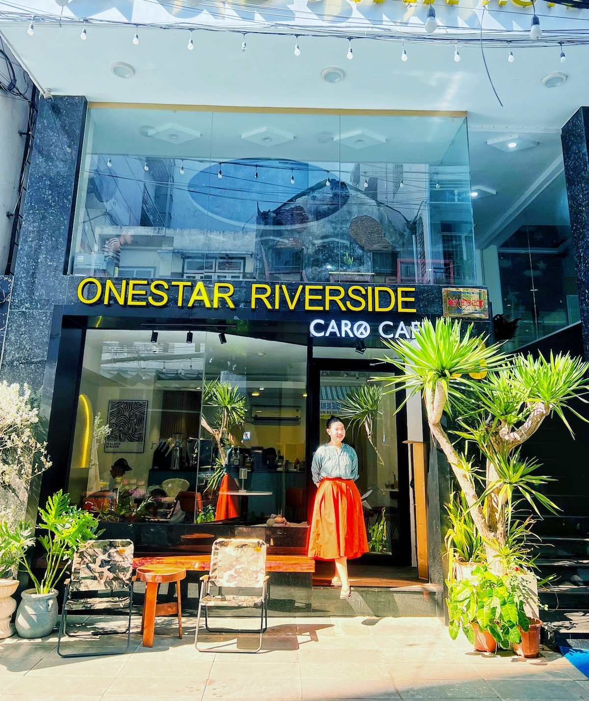 The entrance of Onestar Riverside is captured, showcasing a modern facade with large glass windows. Potted plants flank the entrance, complementing the wooden seating area furnished with patterned chairs. Above, the sign 'ONESTAR RIVERSIDE' is prominently displayed, inviting guests into the space.
