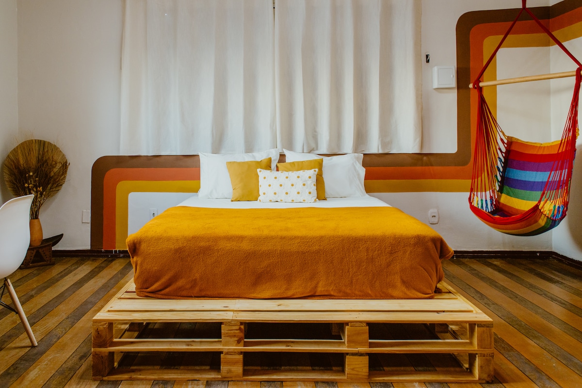 A spacious sleeping area features a wooden platform bed with an orange blanket and decorative pillows. A vibrant, rainbow-colored hammock is suspended nearby, with soft sunlight filtering through sheer curtains. The floor consists of wooden planks, adding warmth to the space.