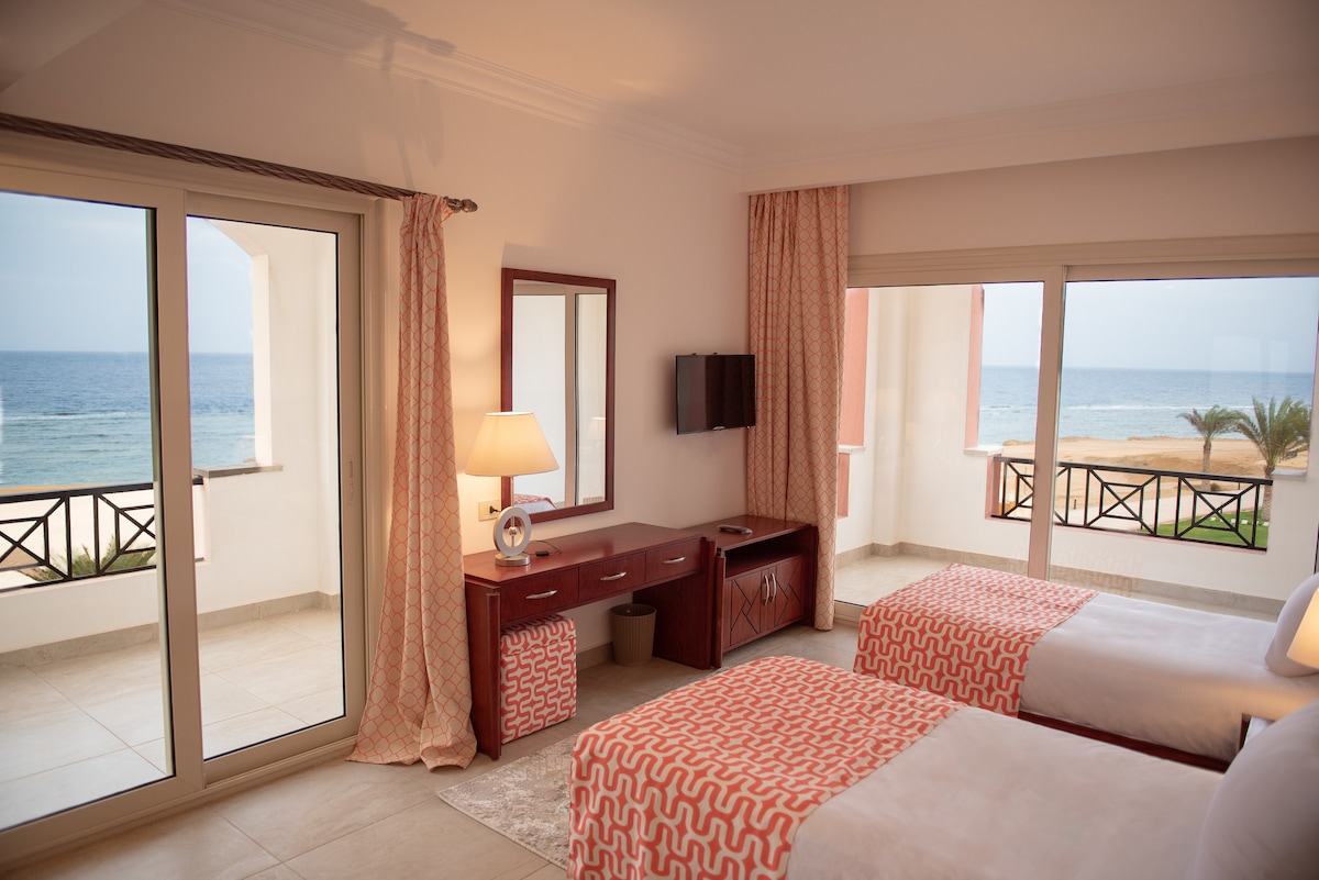 A bright and airy room features two beds adorned with coral-patterned comforters. Large windows frame scenic views of the beachfront and ocean. A wooden dresser and a flat-screen TV complement the space, with natural light illuminating the room throughout.