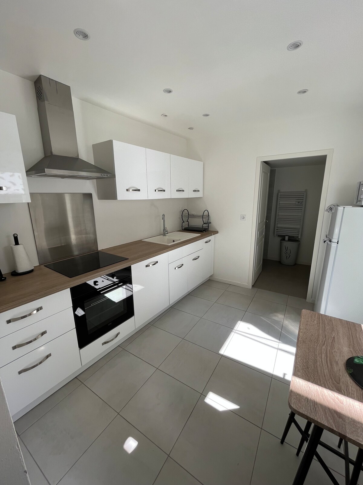 The modern kitchen features clean, white cabinetry and a light-colored countertop. An oven, stove, and sink are integrated into the design, with ample workspace and natural light streaming through a window. A compact dining table is visible, providing convenience for meals.