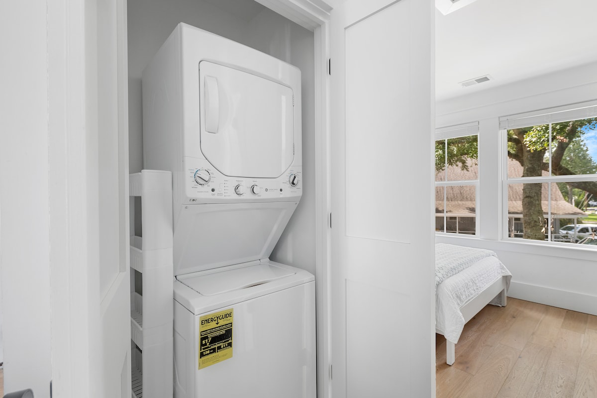 A compact laundry area is displayed, featuring a stacked washer and dryer unit in a bright white finish. The door is open, revealing the appliances inside. Natural light flows in from nearby windows, illuminating the clean, minimalist design of the space.