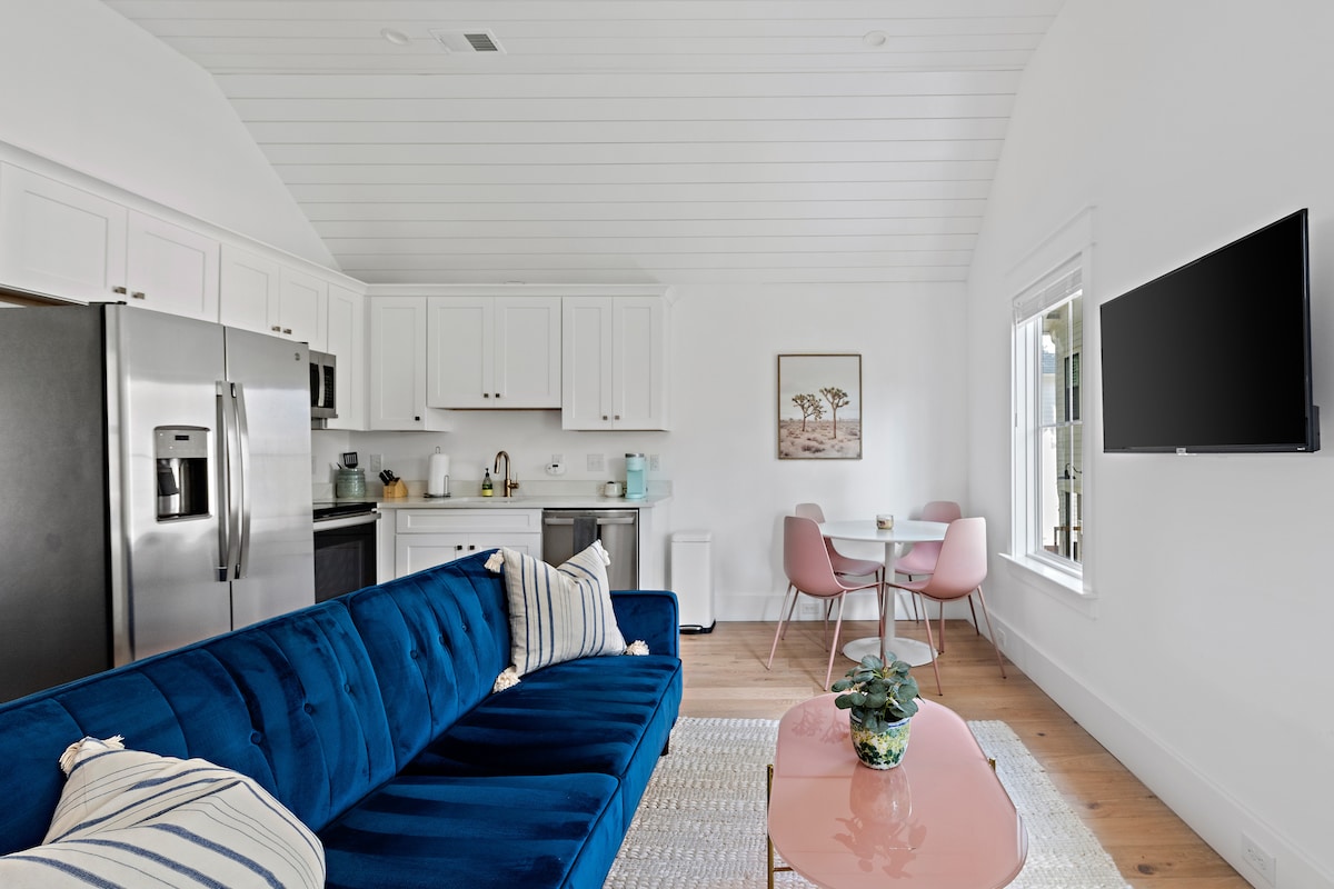 A bright living area features a deep blue sofa with decorative pillows, facing a mounted television. A charming dining set with pink chairs and a round table sits adjacent to the fully equipped kitchen, where stainless steel appliances complement white cabinetry.