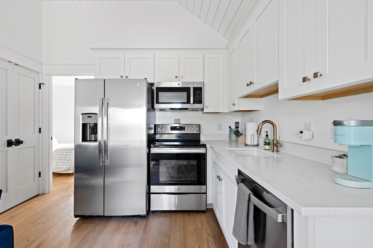 A modern kitchen features stainless steel appliances, including a refrigerator, stove, and microwave. White cabinetry complements the light countertops, and a light blue mixer adds a pop of color. The space is organized with ample counter and storage areas, providing a functional cooking environment.