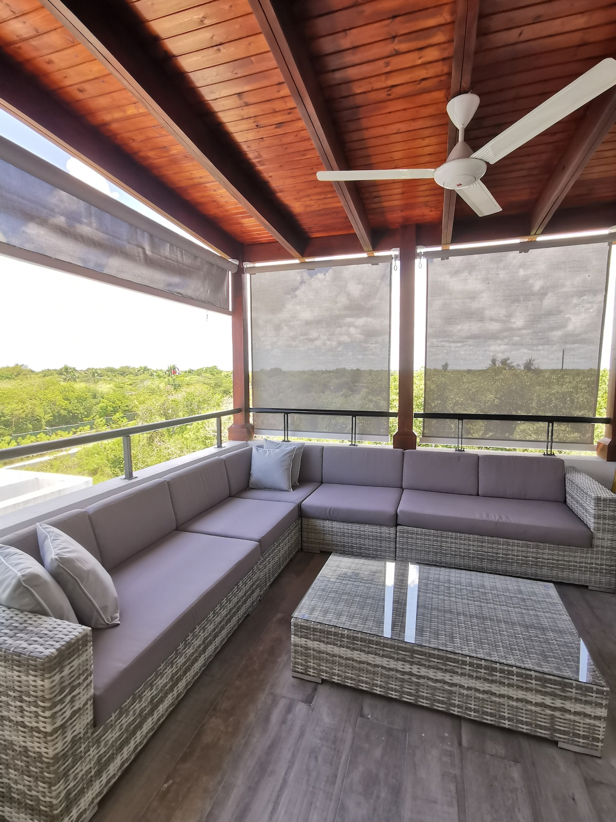 An outdoor balcony features a spacious, sectional seating area with gray cushions, accompanied by a glass-topped coffee table. Adjustable shades provide privacy and shade, while a ceiling fan offers comfort. Lush greenery is visible in the background, enhancing the serene atmosphere.