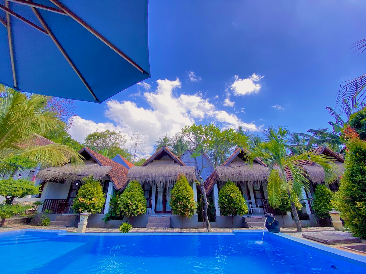 A series of thatched-roof bungalows is arranged around a vibrant blue pool. Lush greenery and palm trees surround the area, while a clear blue sky with scattered clouds provides a serene backdrop.