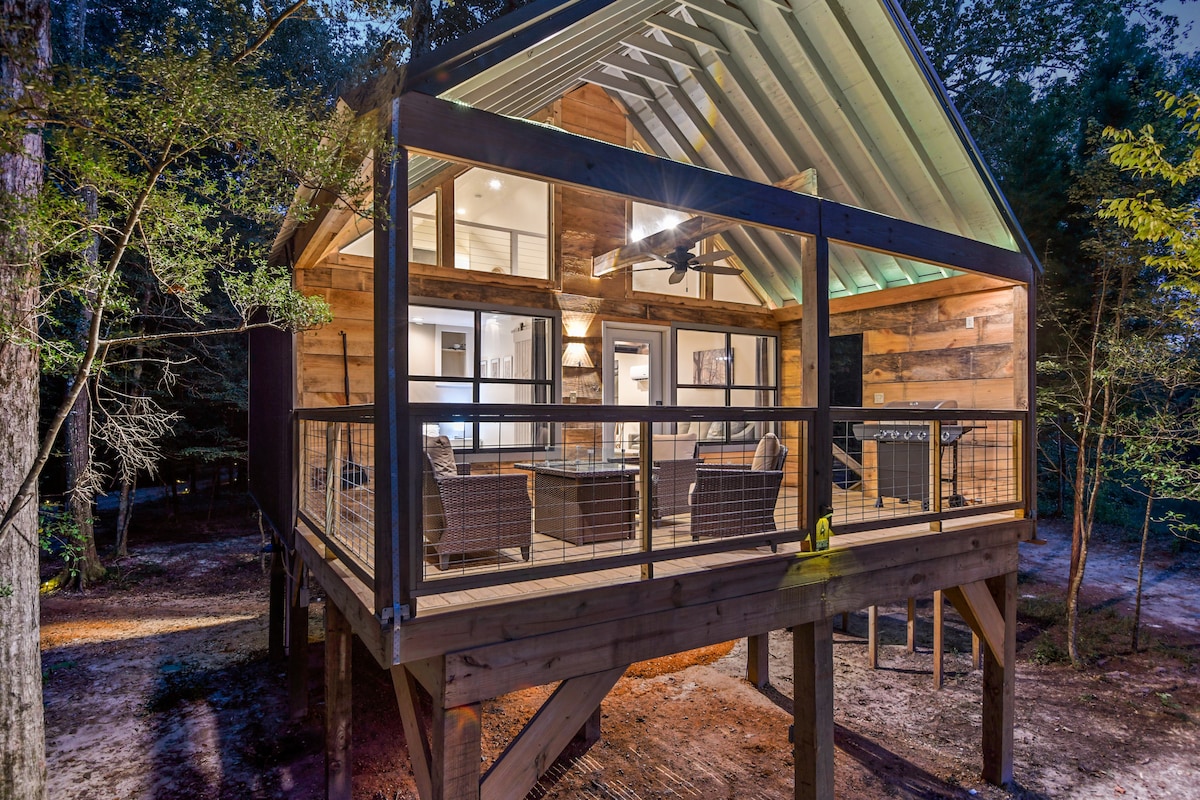 A modern cabin elevated on stilts is surrounded by trees, featuring large glass panels that offer views of the forest. The outdoor deck includes a sitting area with woven chairs. The roof is designed with a distinctive peak, complementing the natural surroundings.
