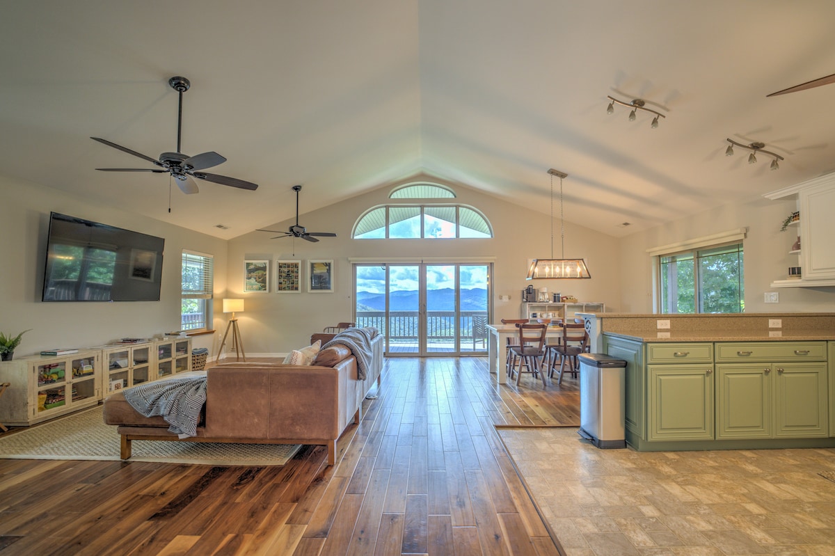 An open living space features large windows that frame mountain views. A comfortable sofa is positioned near a wall-mounted television. Wooden flooring extends through the area, leading to a dining space with a long table, while a kitchen with green cabinetry is visible in the background.