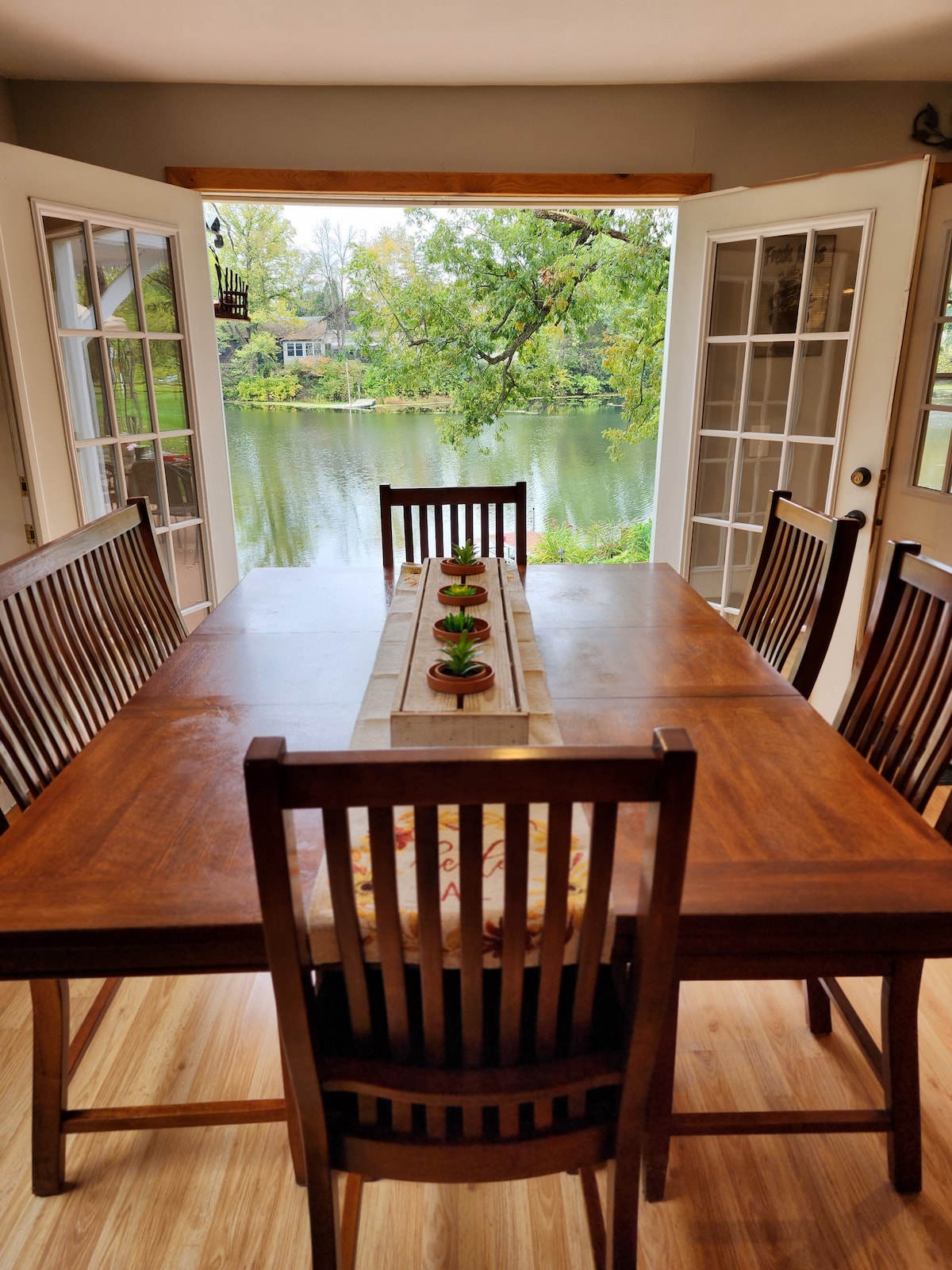 A dining table made of dark wood is set with a long centerpiece, facing large double doors that open to a serene lake view. Surrounding the table are six wooden chairs, creating a comfortable space for gatherings. Natural light is welcomed by the open doors.
