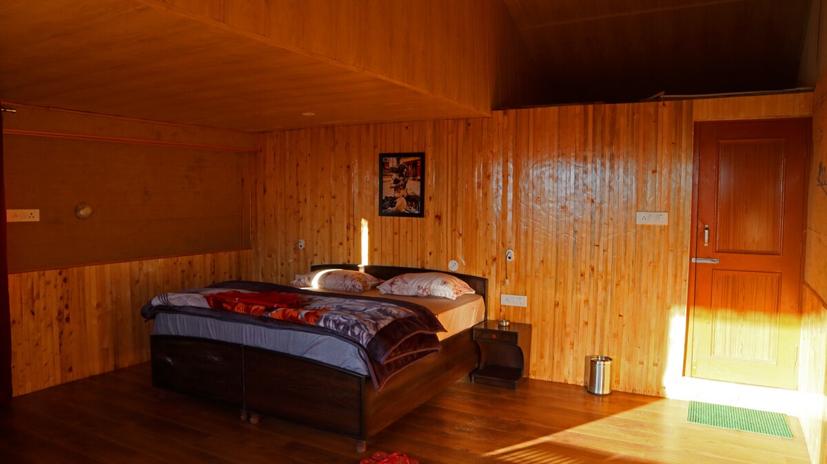 The spacious bedroom features a wooden wall and floor, creating a warm ambience. A large bed is positioned centrally, adorned with folded blankets. Nearby, a wall-mounted light and a nightstand are visible. Light streams in from a window, revealing a welcoming atmosphere.