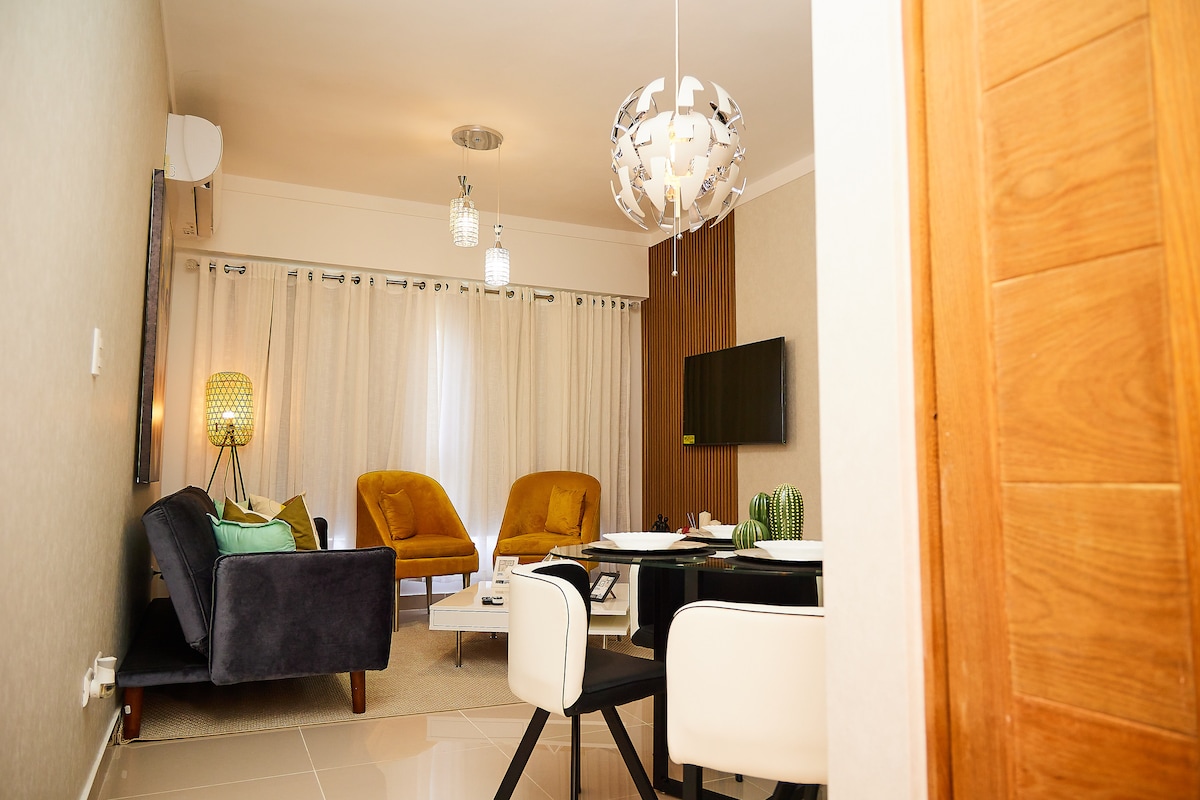 A stylish living area features two yellow armchairs and a dark blue sofa, surrounded by neutral-toned walls. A modern light fixture hangs from the ceiling, and a wall-mounted television is visible. Soft curtains allow natural light to enter the space.