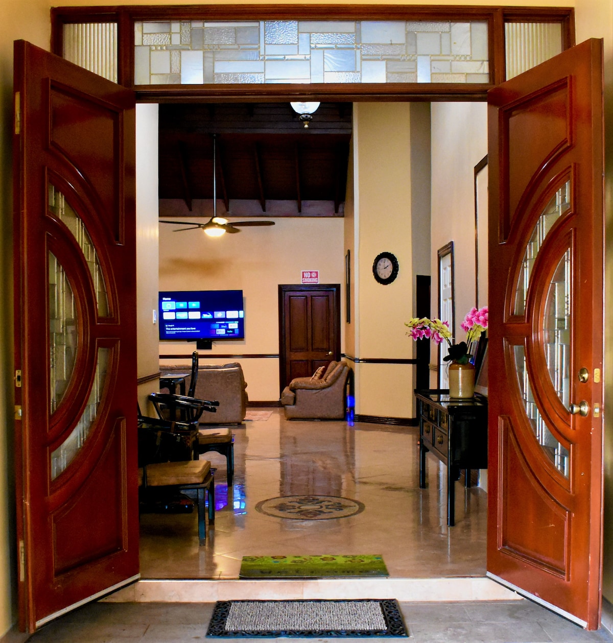 A welcoming entryway features large, ornate double doors, allowing a view into a spacious living area. A sleek television is mounted on the wall, with comfortable seating arranged around it. Decorative plants are situated strategically, enhancing the overall ambiance of the space.