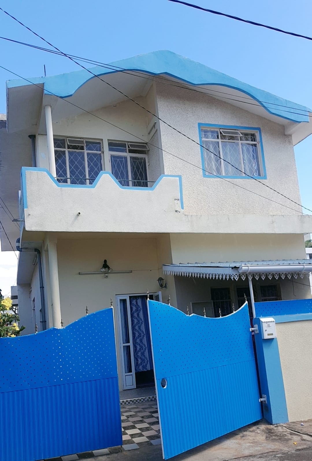 A two-story house is presented, featuring a distinctive blue roof and gate. The entrance is framed by a decorative awning, and large windows allow natural light to illuminate the exterior. The façade displays a blend of subtle architectural design and inviting colors.