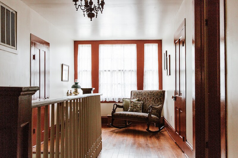 A bright hallway features light wooden floors and white walls. Large windows with sheer curtains provide natural light. A decorative rocking chair with cushions is positioned near the windows, while the hallway is accented by two doors and framed artwork on the walls.