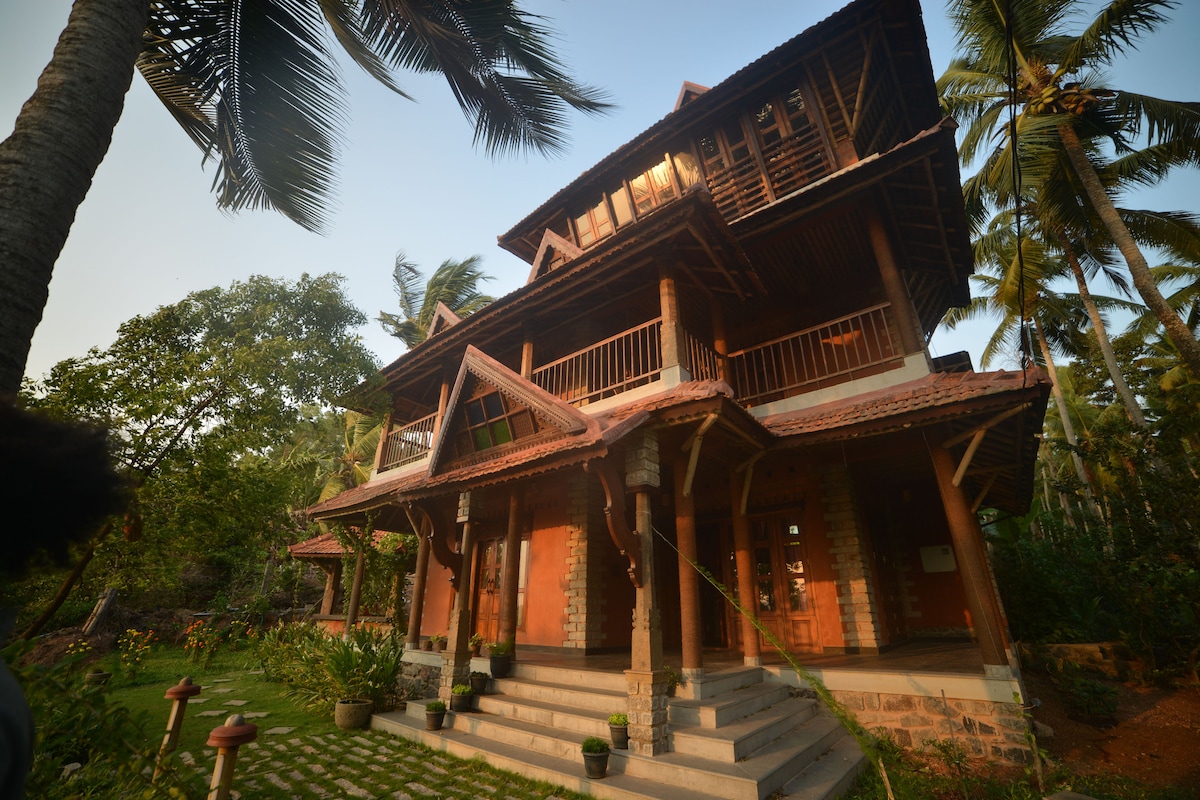 A three-story villa is surrounded by lush greenery, featuring a distinctive architectural style with wooden elements and a terracotta facade. Balconies extend from the upper levels, while stone steps lead up to the entrance. Potted plants and decorative stones line the pathway.