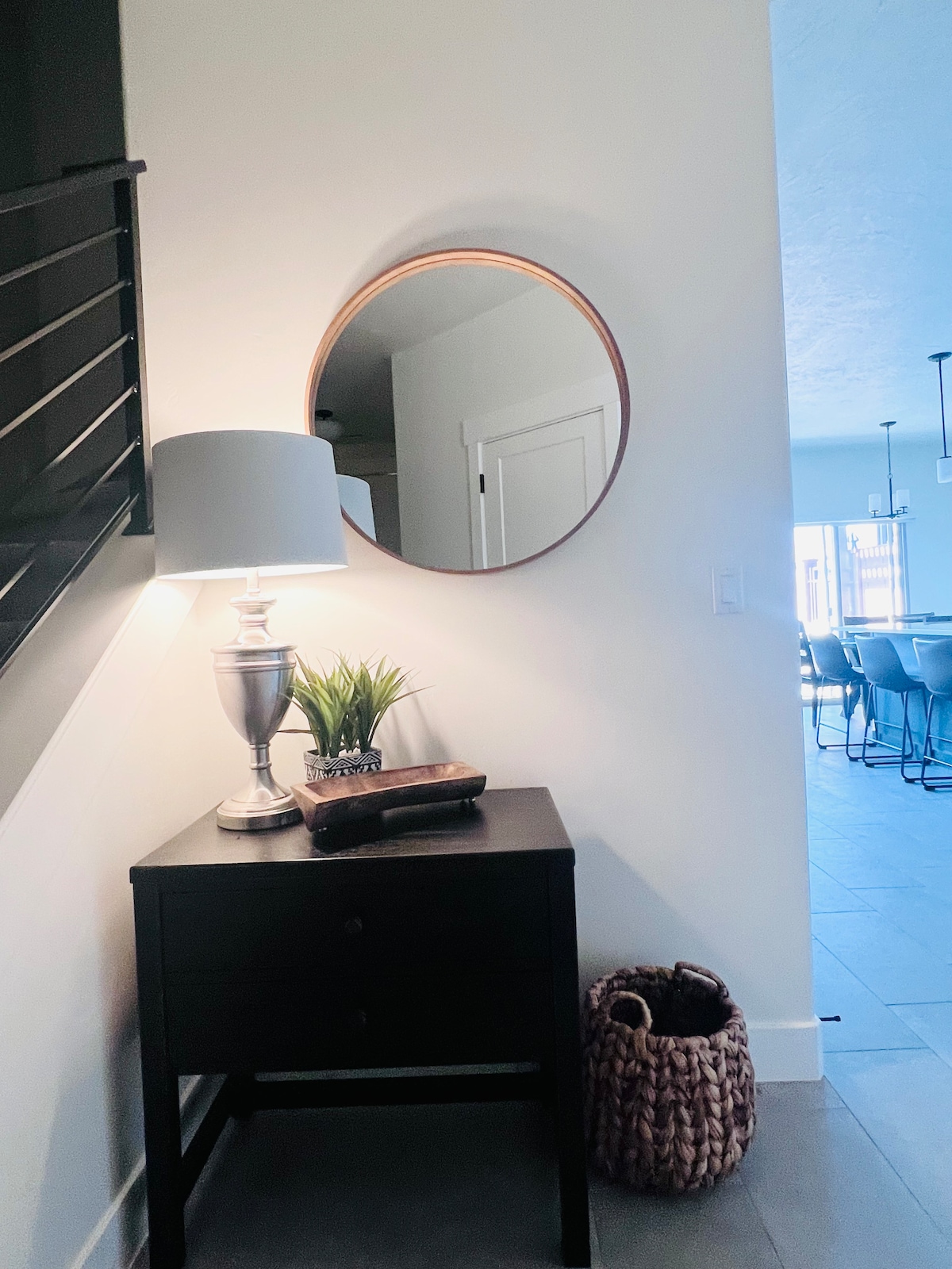 A corner features a wooden side table topped with a silver lamp and a small plant. A round mirror hangs above the table, reflecting the light. A decorative woven basket sits on the floor nearby, with a glimpse of seating and dining areas visible in the background.