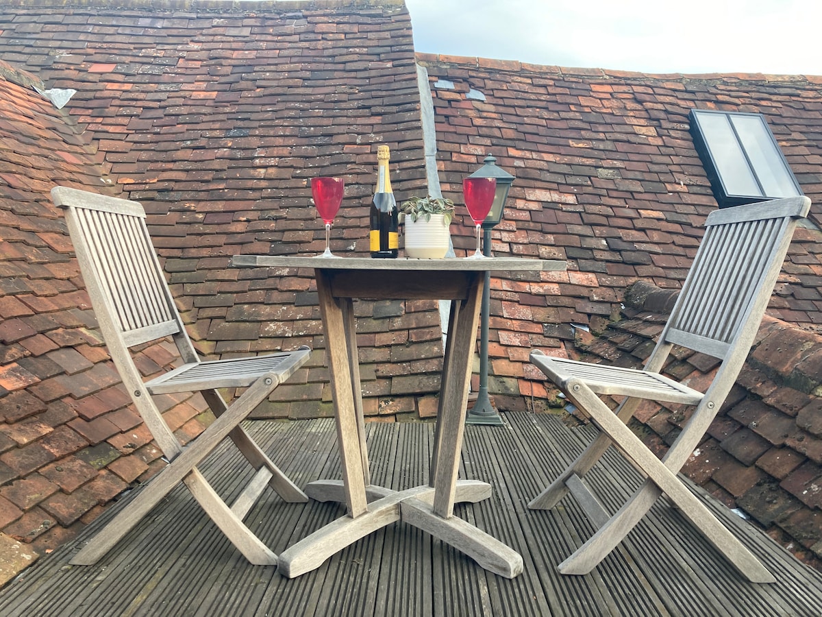 An outdoor dining area is highlighted with a wooden table surrounded by two folding chairs. On the table, a bottle of champagne is placed next to two red glasses and a small potted plant. The backdrop features rustic rooftops.