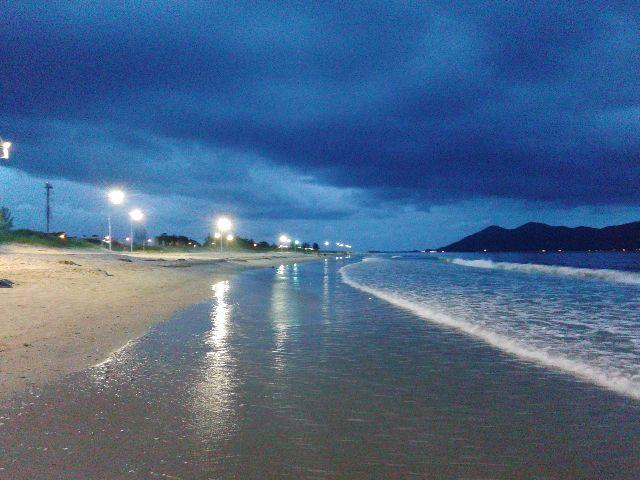 The image captures a serene beach scene at dusk, with gentle waves lapping at the shore. Soft reflections of lights from nearby facilities shimmer on the water's surface. Darkened clouds create a tranquil atmosphere, complementing the expansive sandy beach alongside a distant outline of rolling hills.