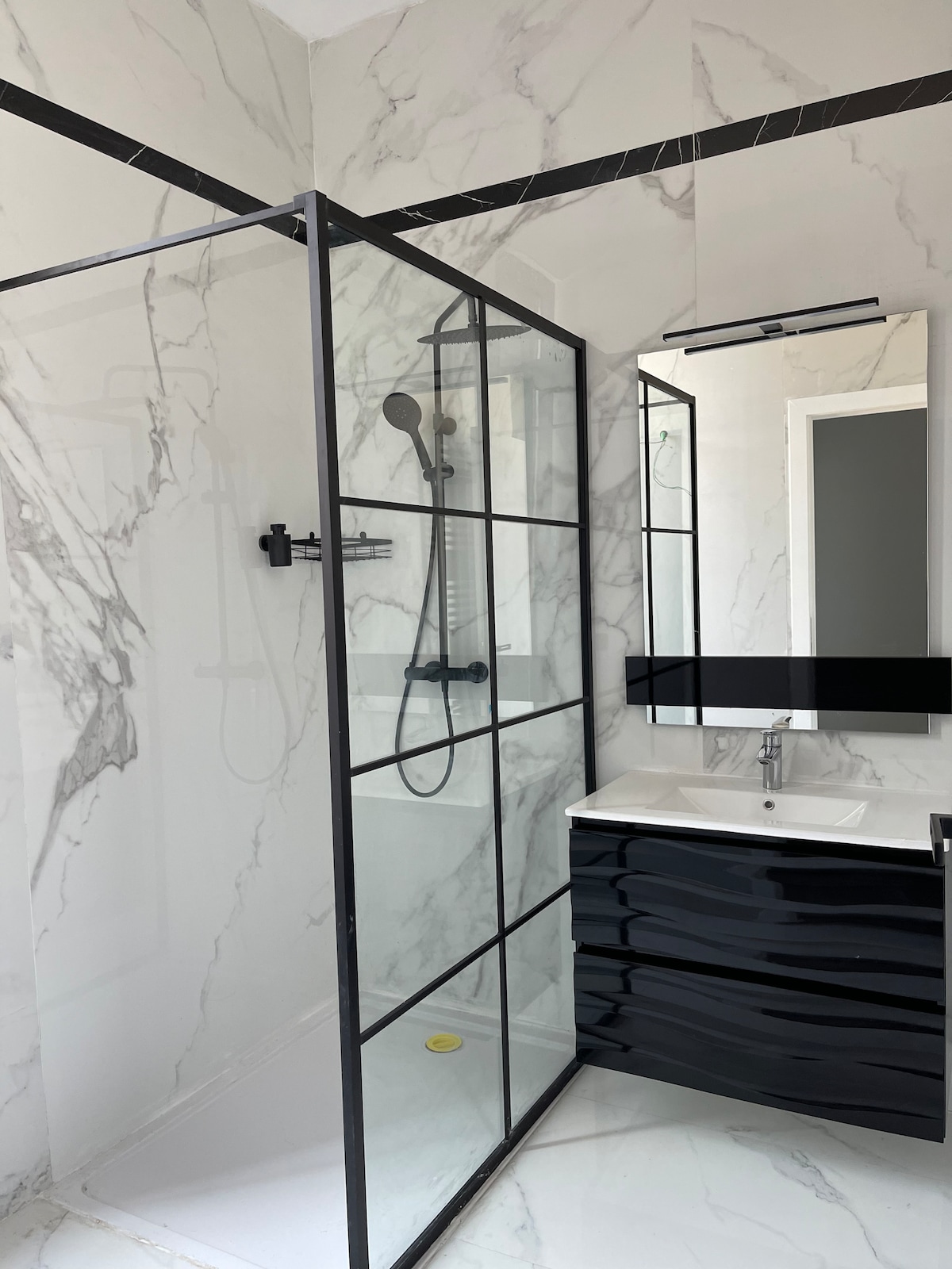 A modern bathroom features a glass-enclosed shower with black framing and a handheld showerhead. A sleek black vanity with a white countertop provides storage. Light-colored marble walls and flooring contribute to a contemporary aesthetic, accented by a large mirror that reflects the space.