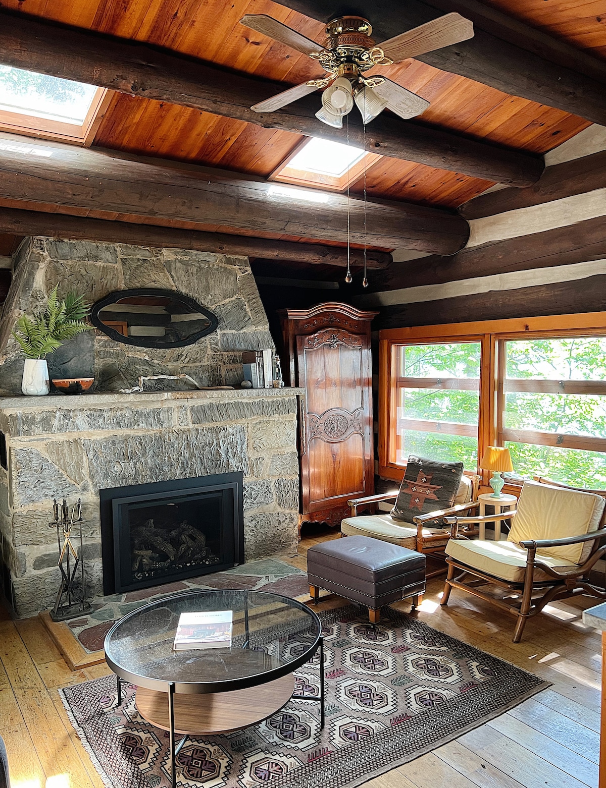The living area features a stone fireplace set against rustic log walls, skylights above, and large windows inviting natural light. A wooden cabinet stands alongside a cozy seating arrangement, including two armchairs and a coffee table placed over a decorative rug.