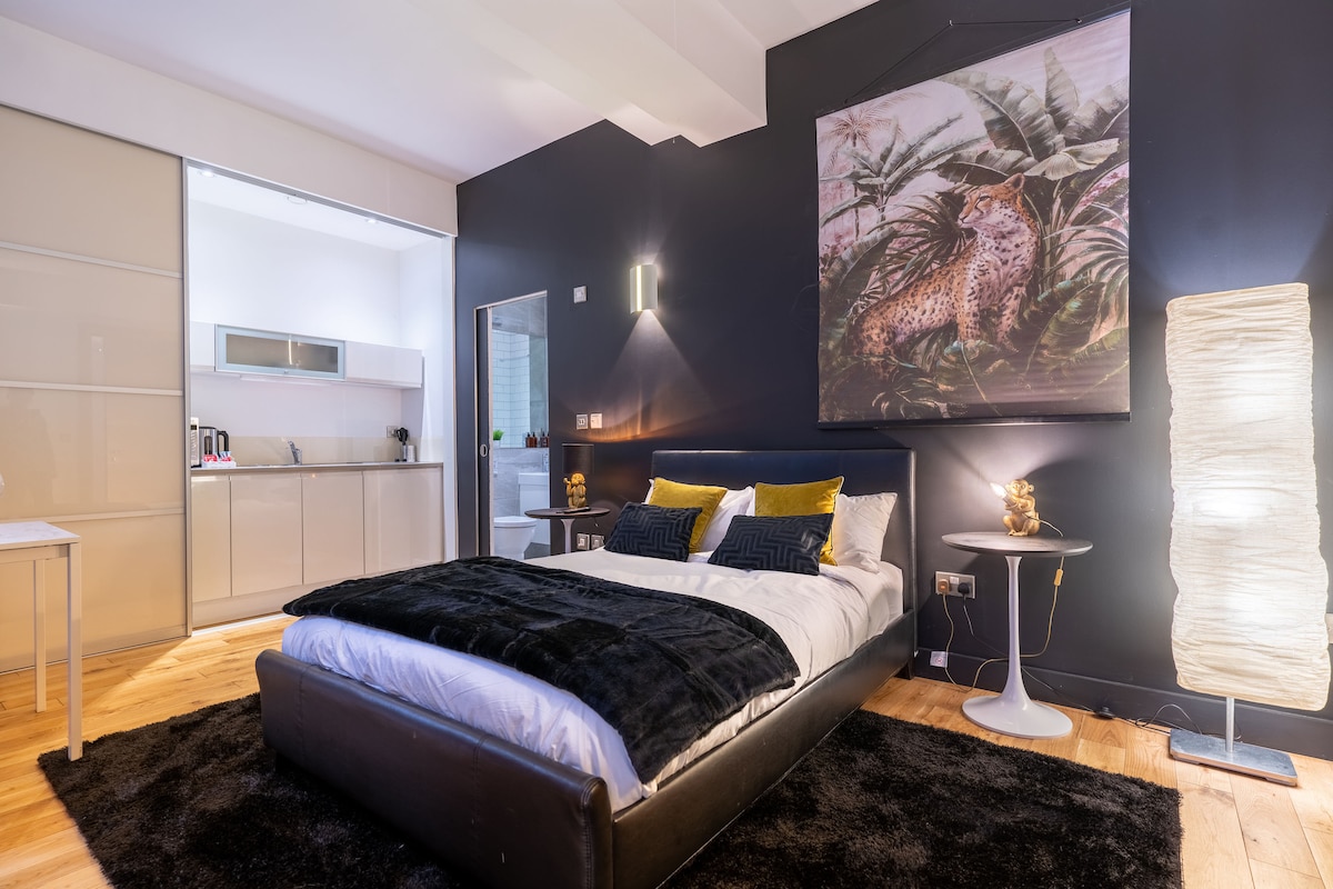 A stylish bedroom features a spacious bed dressed in crisp linens and a dark plush throw. A large piece of wall art depicts tropical foliage. A kitchenette is visible in the background, complemented by modern fixtures and soft lighting. A floor lamp adds gentle illumination.