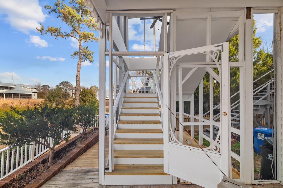 An open entrance leads to a staircase with wooden steps, framed by decorative railings. Natural light illuminates the interior, highlighting the pathway that ascends to the living spaces above. The surrounding greenery offers a serene environment, enhancing the welcoming entryway.