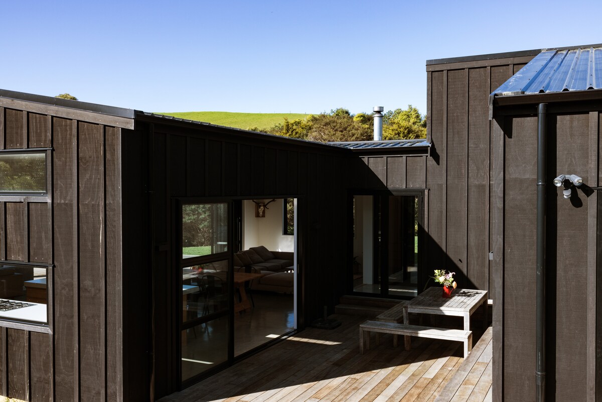 A contemporary home exterior showcases wooden cladding with large glass windows, allowing natural light to fill the interior. A wooden deck features a bench and is bordered by lush greenery, with blue skies visible overhead, creating an inviting outdoor space.