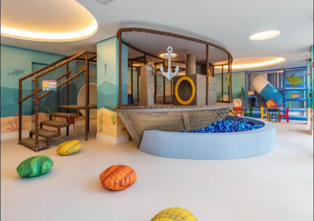 A playful children's area features a large boat structure with climbing stairs and colorful cushions scattered on the floor. Bright murals of aquatic life adorn the walls, creating a fun and engaging environment for kids.
