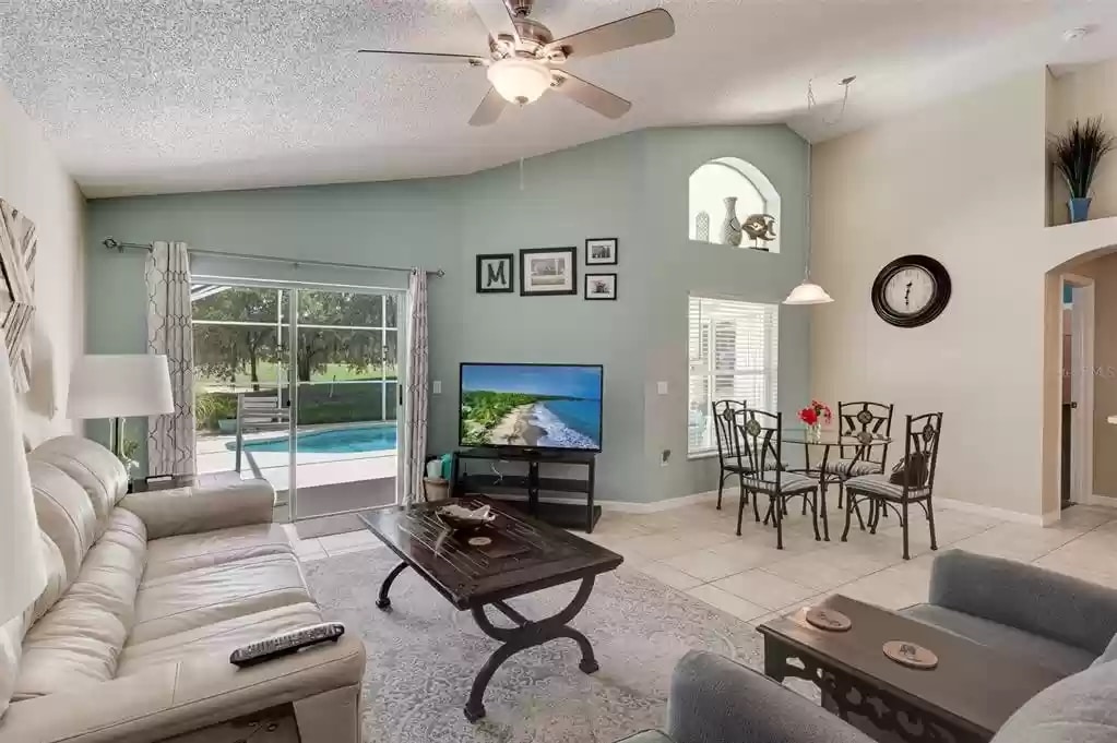 A light-filled living space features a comfortable sofa set, a 60" 4K Smart TV, and a wooden coffee table. A dining area is included with a round table for four. Large windows provide views of the private pool area and allow natural light to enhance the room.
